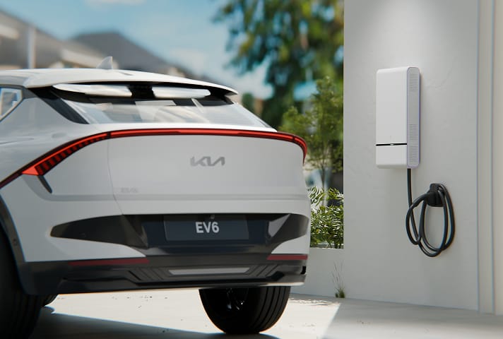 Outdoor shot of the rear of a white Kia EV6 parked next to a vehicle-to-home battery charging station mounted on an exterior white wall, with cable coiled around a hook. Trees and a blue sky are blurred in the backdrop.
