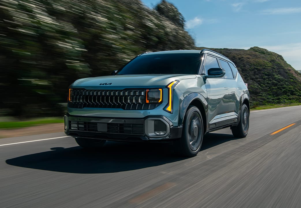 Shot of the 2027 Kia Seltos Hybrid in blue driving quickly through a road surrounded by green hills. The image highlights the compact SUV's bold front grille, tinted windshield, and LED headlights.