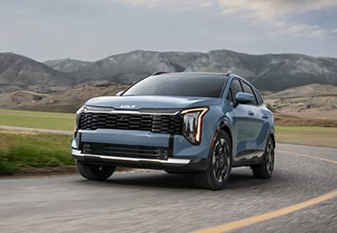 Dynamic shot of the 2027 Sportage Hybrid in blue speeding along a curvy scenic mountain road, featuring a bold front grille, striking LED headlights, and distinctive wheel designs.