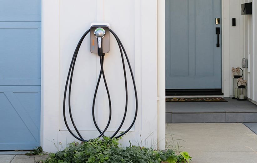 A Level 2 ChargePoint Home Flex hybrid-electric vehicle charging cable and connector, coiled up and attached to the side of a Kia owner's home garage, with shrubs and a blue front door in the background. 
