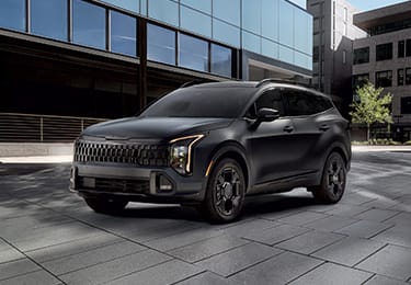 2027 Kia Sportage Plug‑In Hybrid parked in an urban setting, shown from the front with vertical LED headlights, a bold grille design, and modern architectural buildings in the background.